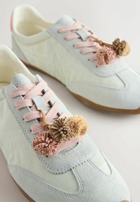 Baskets décontractées blanches et gris clair avec des lacets roses et des pompons décoratifs beige et marron sur un fond clair.
