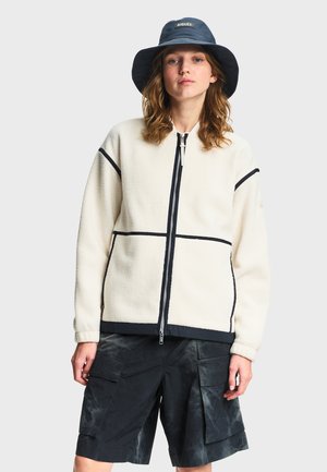 Donna che indossa una giacca in pile bianca con rifiniture nere, pantaloncini cargo neri e un cappello a secchiello blu navy con una toppa logo.