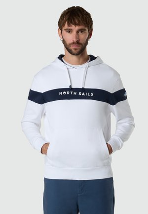 Uomo con felpa bianca con striscia orizzontale blu navy e scritta "NORTH SAILS", mani nelle tasche frontali, pantaloni blu, sfondo neutro.