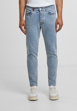 Pantalones de mezclilla ajustados de color azul claro, con diseño de cinco bolsillos, costuras rojas en contraste y textura envejecida. Combinados con zapatillas blancas.