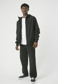 Schwarze leichte Jacke mit Kapuze, Reißverschluss, elastischen Bündchen, getragen über einem weißen Hemd und kombiniert mit schwarzen Hosen und schwarzen Sneakers.