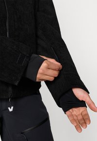 Černá vroubkovaná fleecová bunda se zipovou kapsou, disponující integrovanými černými elastickými rukávy a uzávěry na suchý zip.