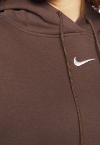 Ruskea huppari, jossa on teksturoitu pinta, hopeinen Nike-logo ja kiristysnauhat, korostaen sen rentoa muotoilua.
