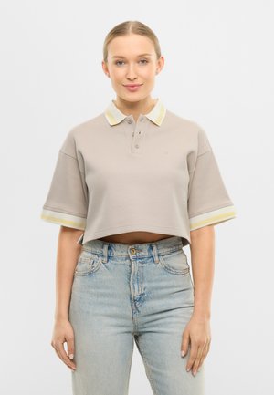 Nuori nainen, jolla on beige lyhyt poolopaita, jossa on keltaiset ja valkoiset raidalliset hihat, ja vaalean siniset korkeavyötäröiset farkut, seisomassa valkoista taustaa vasten.