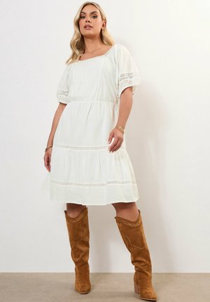 Robe en coton blanc avec des manches bouffantes courtes, ornée de dentelle et d'un ourlet à plusieurs niveaux. Associée à des bottes en daim hautes jusqu'aux genoux de couleur beige.