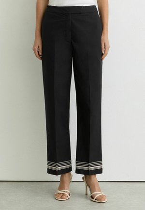 Fekete egyenes szárú nadrág fehér vízszintes csíkokkal a szegély közelében, bézs magas sarkú szandállal viselve.