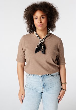 Vrouw met krullend haar die een bruin kort mouwtopje draagt, lichtblauwe spijkerbroek, sjaal met luipaardprint, creolen en armbanden, staand tegen een witte achtergrond.