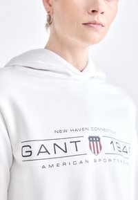Hvit hettegenser med et trykk på forsiden som viser "GANT," "New Haven Connecticut," og et emblem med stjerner og striper. Myk tekstur.