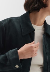Personne portant une veste en velours côtelé foncée par-dessus une chemise claire côtelée, tenant la fermeture éclair de la veste près du col avec la main droite.