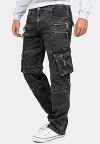 Pantaloni cargo in denim nero con molteplici tasche con cerniera, dettagli trapuntati e vestibilità affusolata. Abbinati a sneakers bianche e una felpa grigia.