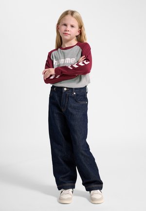 Blond barn står med armene over kors iført grå og vinrød langærmet trøje, mørkeblå jeans og hvide sneakers mod hvid baggrund.