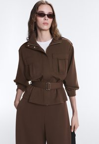Femme portant une veste marron ceinturée avec de grandes poches, un pantalon assorti, des lunettes de soleil et une chemise blanche en dessous, posant devant un fond uni.