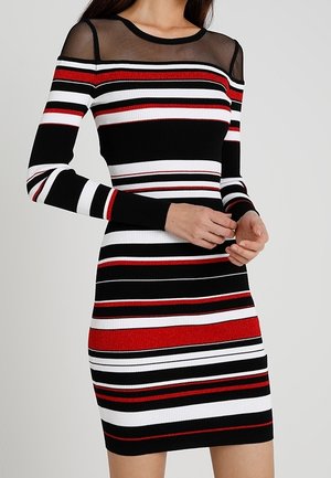Femme portant une robe longue jusqu'aux genoux, à manches longues, avec des rayures horizontales noires, blanches et rouges et un empiècement en maille noire transparente au niveau de l'encolure.