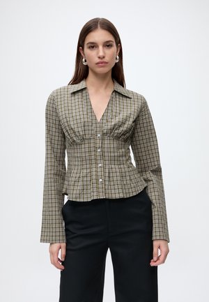 Femme portant un chemisier cintré à carreaux verts et blancs, à boutons, manches longues, avec un pantalon noir, debout devant un fond uni.