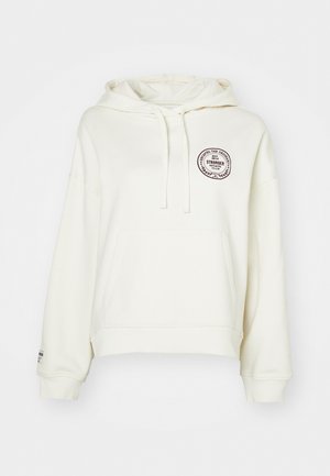 Hanorac de culoare crem, cu o croială lejeră, glugă cu șnur și buzunar frontal. Prezintă o patch logo rotund de culoare maro închis pe piept.