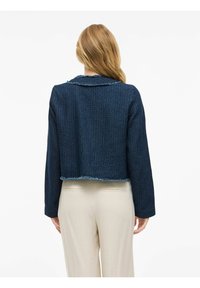 Žena s dugom plavom kosom koja nosi skraćeni teksturirani tamnoplavi sako i svijetlosmeđe hlače, stoji okrenuta leđima prema jednostavnoj pozadini.