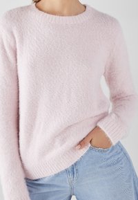Pull léger en rose fluffy avec un col rond et des poignets côtelés. Porté sur un jean bleu, affichant une apparence douce et texturée.