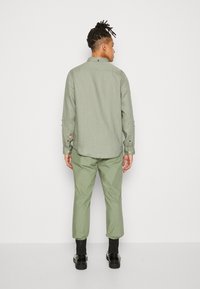 Camisa de manga larga verde clara con collar estándar, puños abotonados y dobladillo curvado; llevada con pantalones verdes a juego y zapatos negros.