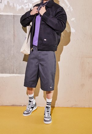 Czarna kurtka, fioletowa koszula, szare szorty, czarno-białe skarpetki w paski, szare sneakersy za kostkę. Bawełniana torba na ramieniu. Dyskretne logo.