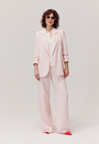 Jasnoróżowa marynarka i spodnie z szerokimi nogawkami, charakteryzujące się gładką teksturą i dopasowanym krojem. Biała bluzka pod spodem, czerwony akcent na butach.