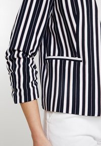 Marineblauw-wit gestreepte blazer met een getailleerd ontwerp, voorzien van dunne roze strepen, een enkele zak en driekwart mouwen.