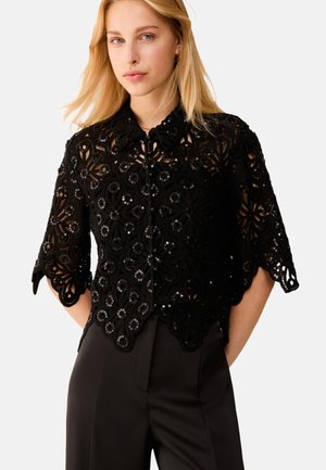 Donna bionda che indossa una camicetta nera in pizzo con maniche corte, paillettes e bordi scallopati, abbinata a pantaloni neri a vita alta.