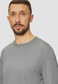 Graues Langarmshirt mit rundem Ausschnitt, aus weichem Stoff gefertigt, mit kleinem Logo auf der Brust. Das Design ist minimalistisch und figurbetont.