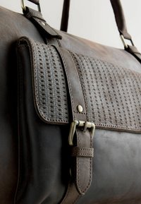 Bruine leren tas met een gestructureerde grijze geweven klep, koperen hardware en een gladde afwerking. Inclusief een gesp en riem voor sluiting.