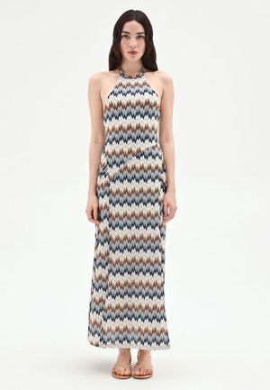 Femme aux longs cheveux foncés portant une robe longue sans manches à col halter avec un motif zigzag marron, bleu et blanc, debout sur fond blanc.