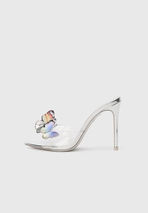 Sandal transparente de tacón alto con un acento metálico en plata y un detalle de mariposa colorida en la tira del dedo, mostrando texturas suaves.