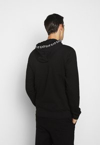 Svart hoodies med vit logotyp tryckt längs huvan, har långa ärmar och en avslappnad passform.