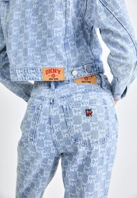 Denimjacka och byxor i ljusblått, med en upprepande logotypmönster. Röda varumärkesetiketter och svarta broderade detaljer är synliga.