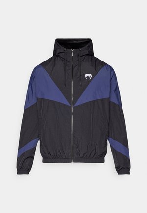 Black and dark blue zip-up windbreaker with hood, elastic cuffs, and a white fang logo on the chest, laid flat on a white background.