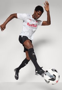 Hvid sportsjersey med "Red Bull" logo, sorte shorts, sorte fodboldsokker og sorte støvler; spiller der sparker til en sort og hvid fodbold.
