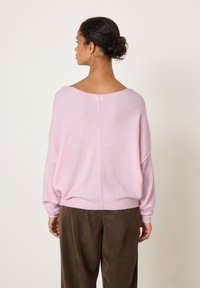 Moteris su tamsiais plaukais surištais kuodu, dėvi auksinius lankinius auskarus, laisvą šviesiai rožinės spalvos megztinį ir rudus kordurojo kelnes, žiūrinti į šalį ant vienspalvio fono.