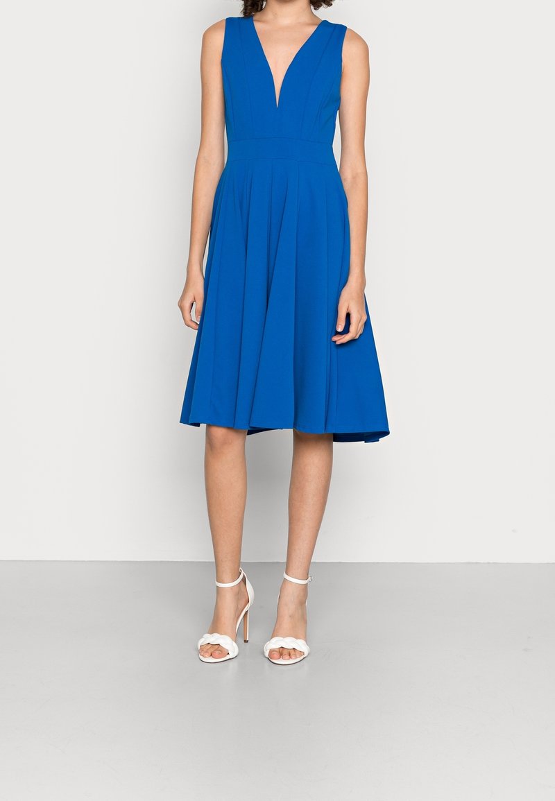 Robe sans manches bleu roi longueur genou avec un profond décolleté en V et une jupe plissée, portée avec des sandales blanches à talons hauts.