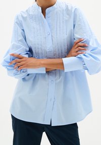 Personne portant une chemise plissée bleu clair boutonnée avec des manches ballon et un pantalon foncé, les bras croisés sur la poitrine.