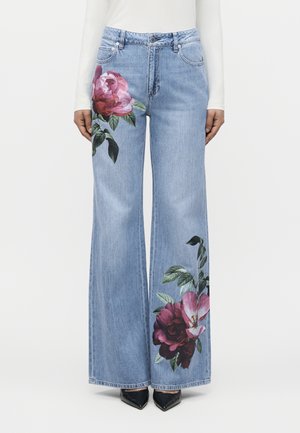 Svetlomodré rifle s širokými nohavicami a veľkými ružovými a červenými kvetinovými vzormi na ľavom stehne a pravom dolnom lýtku, nosené s čiernymi špicatými topánkami.