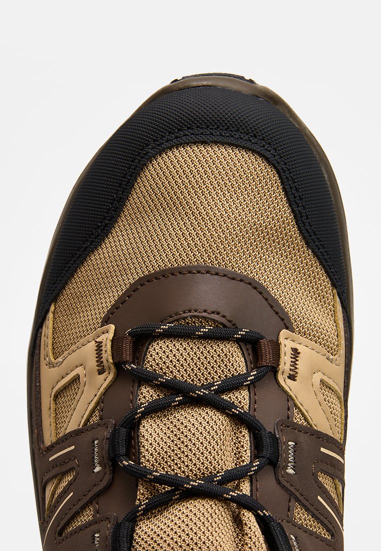 Zapatilla deportiva marrón y beige con parte superior de malla, tapa de goma oscura en la punta y cordones negros. Presenta ojales reforzados y acentos texturizados.