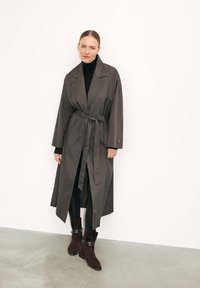 Hosszú, sötétszürke trench coat sz�éles hajtókákkal, övvel a derékon és oldalsó hasítékokkal, fekete, testhezálló nadrággal és barna bokacsizmával párosítva.