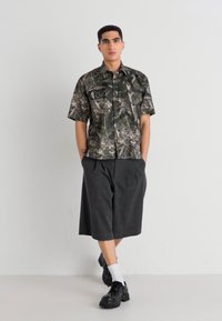 Cămașă cu mâneci scurte și model de camuflaj, asortată cu pantaloni scurți încreți până la genunchi de culoare închisă și pantofi masivi negri, cu șosete albe.