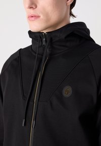 Čierna mikina s kapucňou, ktorá má vysoký golier, predný zips a zlaté akcenty. Výrazné detaily zahŕňajú odznak s logom a textúrovaný materiál.