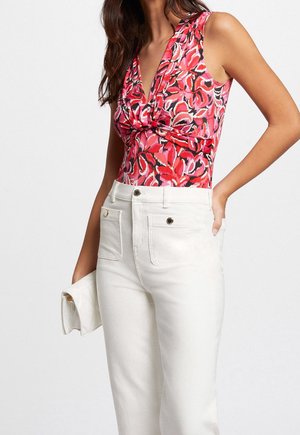 Femme portant un haut sans manches rose à motifs avec un détail noué, et un pantalon blanc taille haute, tenant une pochette blanche dans la main gauche.