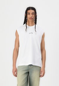 Hvit ermeløs T-skjorte med rund hals, med en liten trykt logo i svart på brystet. Stoffet virker glatt og lett.