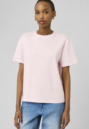 Jeune femme portant un t-shirt rose clair à manches courtes, un jean bleu, des boucles d'oreilles en or en forme d'anneau et des bracelets, debout devant un fond gris.