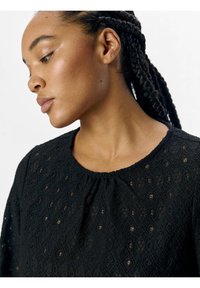 Black lace blouse featuring a round neckline, textured fabric with geometric patterns, and a loose fit with gathered detailing at the neck.