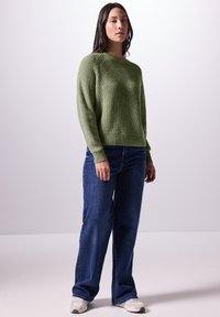 Maglione lavorato a maglia verde con texture a coste, scollo tondo e maniche lunghe, abbinato a jeans blu larghi e sneakers grigie.