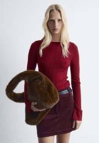 Rotes geripptes Langarmoberteil, kombiniert mit einem bordeauxfarbenen Cordrock, hält eine braune Tasche aus Kunstfell mit einem geschwungenen Griff. Schlichter Hintergrund.