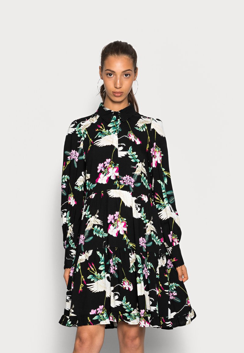 Vestido preto com estampado floral apresentando gruas e orquídeas, mangas compridas e saia em camadas. Detalhes de gola e botões realçam o design.