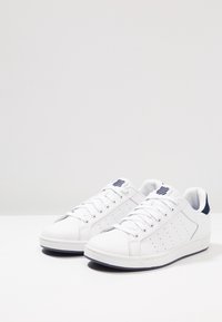 Bílé tenisky vyrobené z syntetického materiálu s námořnickými akcenty. Mají perforované boky, šněrování a plochou podrážku. Klasický design nízkého střihu.